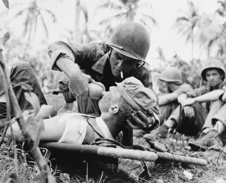 Soldier gives water to wounded comrade on stretcher.